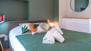 A neatly made bed with a green blanket and white pillows, a bedside table with a lamp, and a round mirror on the wall. - Villa du Port 2 - Appt vue port (La Trinite-sur-Mer)
