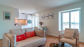 A modern living room with a beige sofa, a red pillow, and a wooden coffee table. There is a dining area with a table and chairs, and a kitchen in the background. - Villa du Port 2 - Appt vue port (La Trinite-sur-Mer)