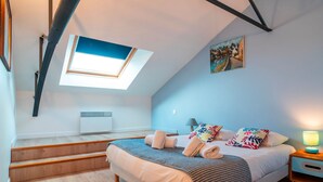 A bedroom with a skylight, a bed with multiple pillows and a blanket, a bedside table with a lamp, and a painting on the wall. - L'Atelier Port Fétan - Vue mer (Locmariaquer)