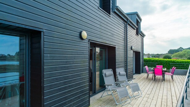 A modern building with a wooden deck, outdoor seating, and a large window. - L'Atelier Port Fétan - Vue mer (Locmariaquer)