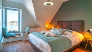 A modern bedroom with a large bed, a view of the city through a window, and a built-in bench with seating. - Villa du Port 6 - Sublime appt vue vieux port (La Trinite-sur-Mer)