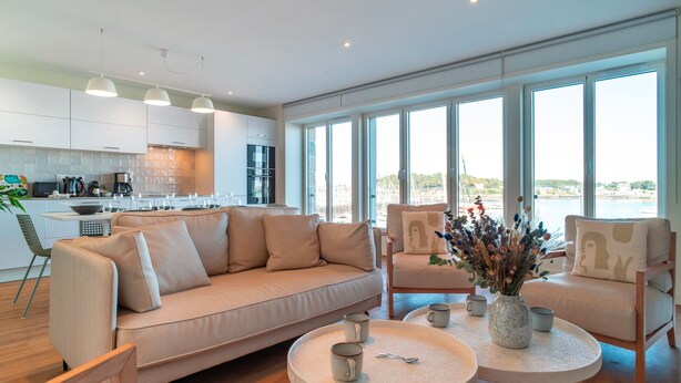 A modern living room with a beige sofa, two armchairs, and a coffee table. There is a kitchen in the background with white cabinets and a large window. - Villa du Port 6 - Sublime appt vue vieux port (La Trinite-sur-Mer)