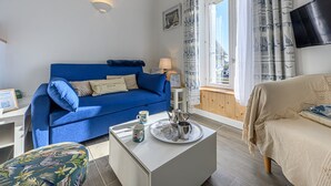 Una sala moderna con un sofá azul, una mesa de centro blanca con tetera y taza, un sofá color beige y una ventana con cortinas.