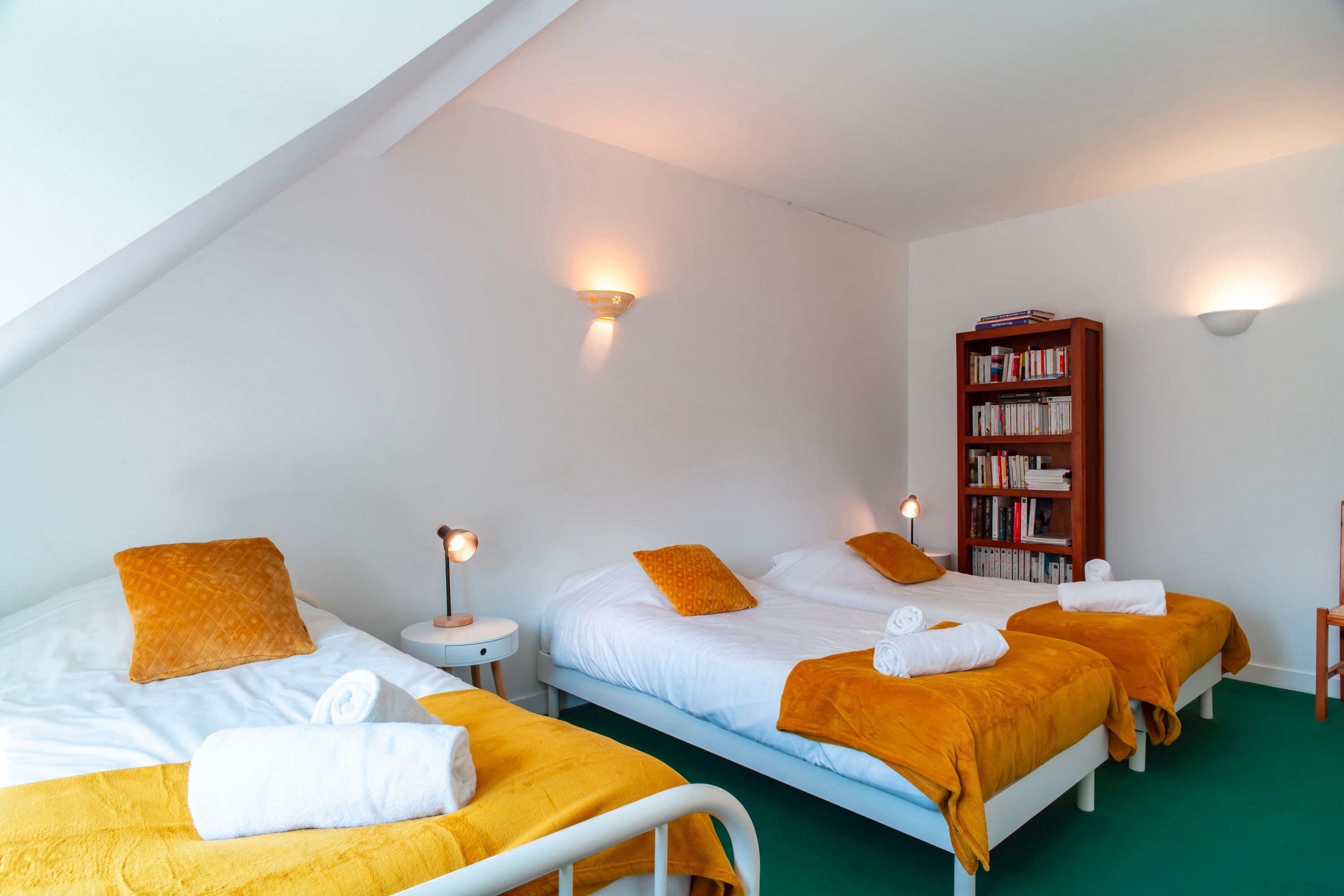 A hotel room with two beds, a bookshelf, and a small table with a lamp.