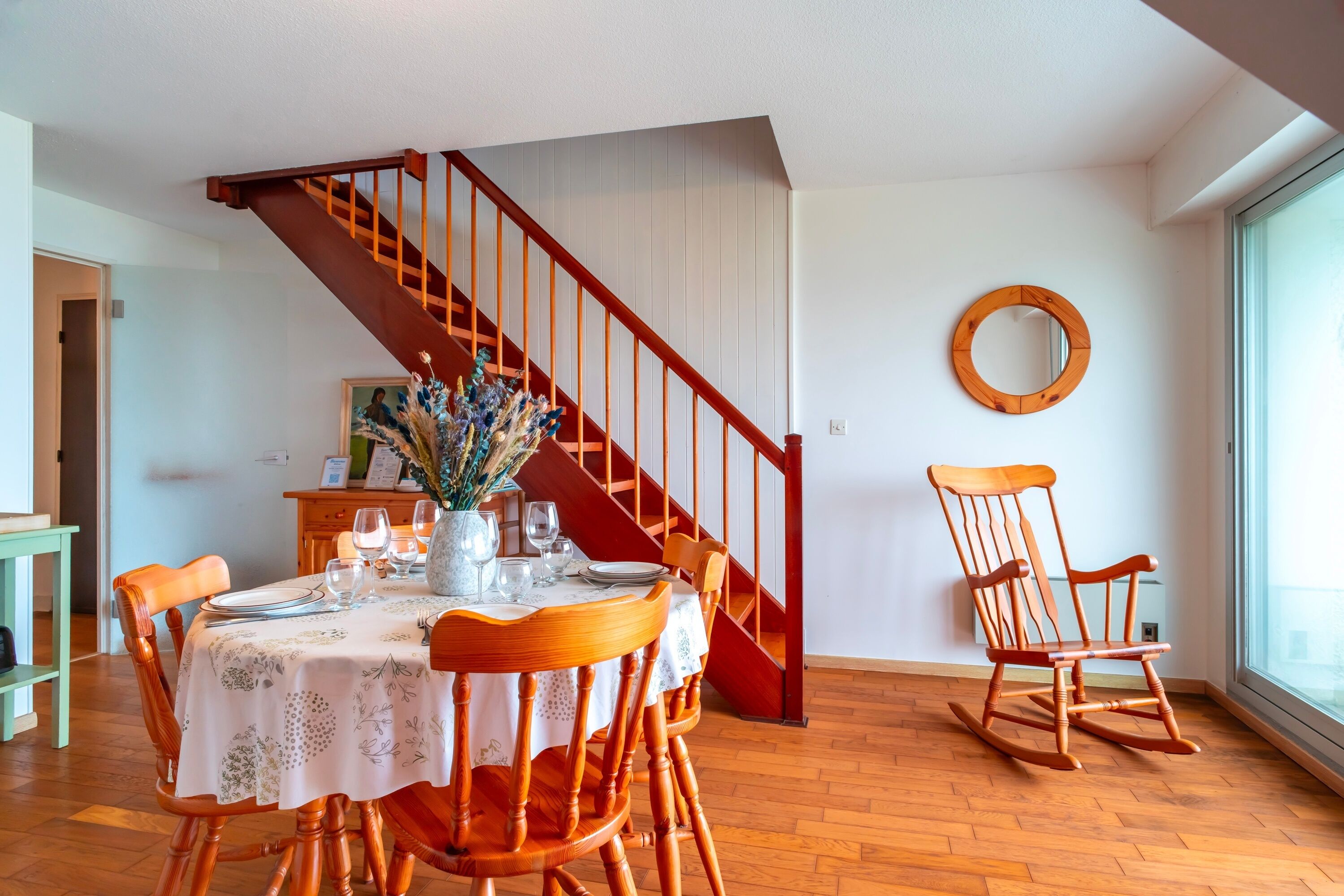 A dining area with a wooden staircase, a dining table set with plates, glasses, and a vase of flowers.
