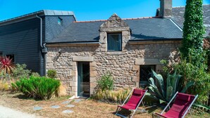 Une maison en pierre avec un toit sombre, une petite fenêtre et une porte. Il y a deux chaises rouges dans le jardin.
