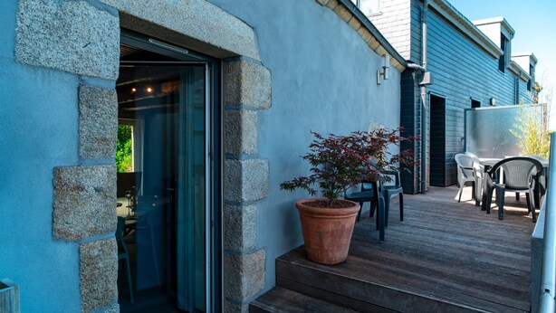 Une façade en pierre, une terrasse en bois, un pot de fleurs et du mobilier d’extérieur.