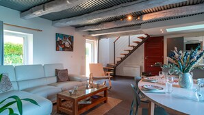 Un salon moderne avec un canapé blanc, une table basse en bois et un coin repas avec une table blanche dressée pour quatre personnes. Un escalier mène à une autre pièce.