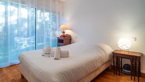 Un dormitorio con una cama grande, una mesita de noche con lámpara y vistas a través de una ventana con cortinas translúcidas.