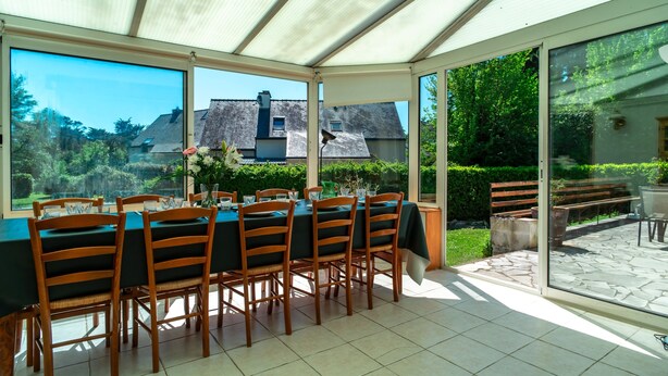 A covered patio with a dining table set for six, large windows, and a view of a house and garden.