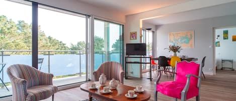 A modern living room with a large glass door leading to a balcony, a dining area with a table set for four, and a bright, airy space with a large window.