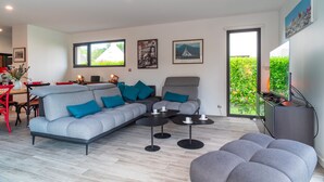 A modern living room with a grey sofa, a grey armchair, and a grey ottoman. There are two black coffee tables and a dining area with red chairs. The room has large windows with a view of greenery outside. - Ti Marsouin - Jolie maison avec extérieur (La Trinite-sur-Mer)