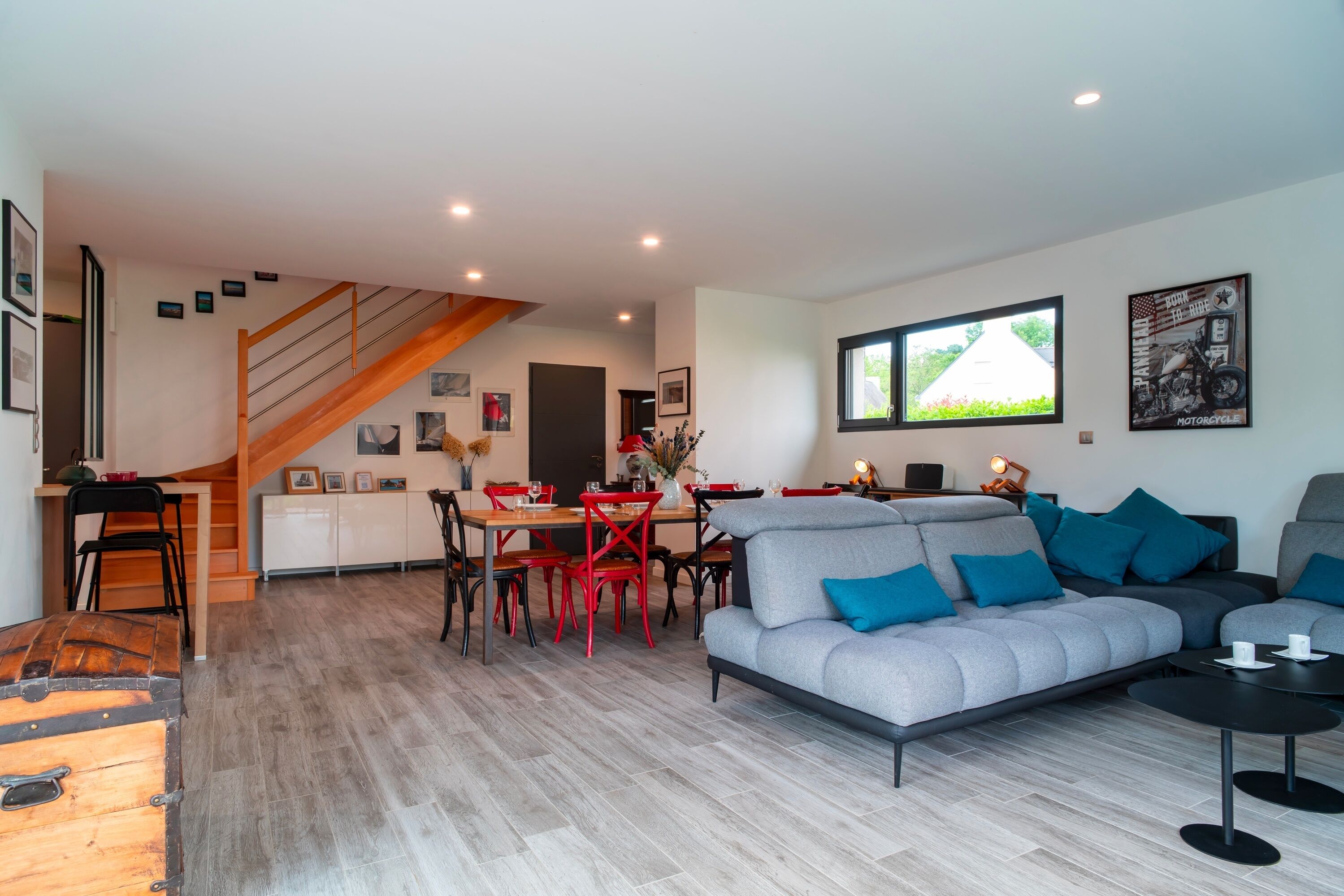 Un salon moderne avec un canapé gris, des fauteuils rouges, une table à manger et un escalier menant à un autre niveau.