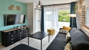 Ein modernes Wohnzimmer mit Flachbildfernseher, einem schwarzen Sofa, einem Holztisch, einer schwarzen TV-Konsole mit Schubladen und Blick ins Freie durch eine Glastür.