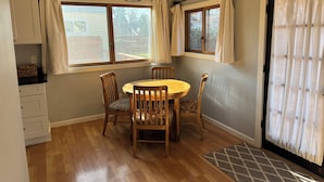 Un coin repas avec une table ronde en bois et quatre chaises, deux fenêtres avec des rideaux, un luminaire au plafond et un plancher en bois.