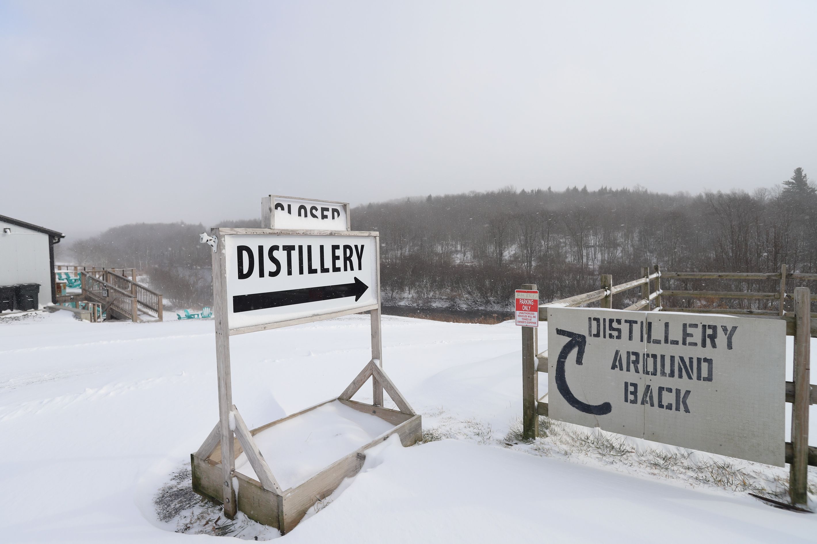 Une enseigne représentant une distillerie, recouverte de neige, avec une flèche indiquant la direction de la distillerie, et une autre enseigne précisant que la distillerie se trouve à l’arrière.
