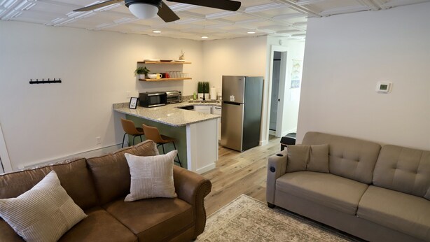 Een moderne woonkamer met een bruine leren bank en een beige bank, een keuken met een roestvrijstalen koelkast, en een plafondventilator.