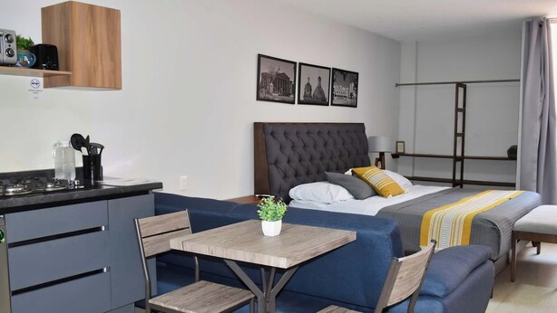 Une chambre d’hôtel compacte comprenant un coin cuisine, un lit avec une tête de lit, une petite table avec des chaises et une plante en pot.
