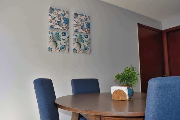A round wooden table with four blue upholstered chairs, a potted plant, and two floral wall art pieces.