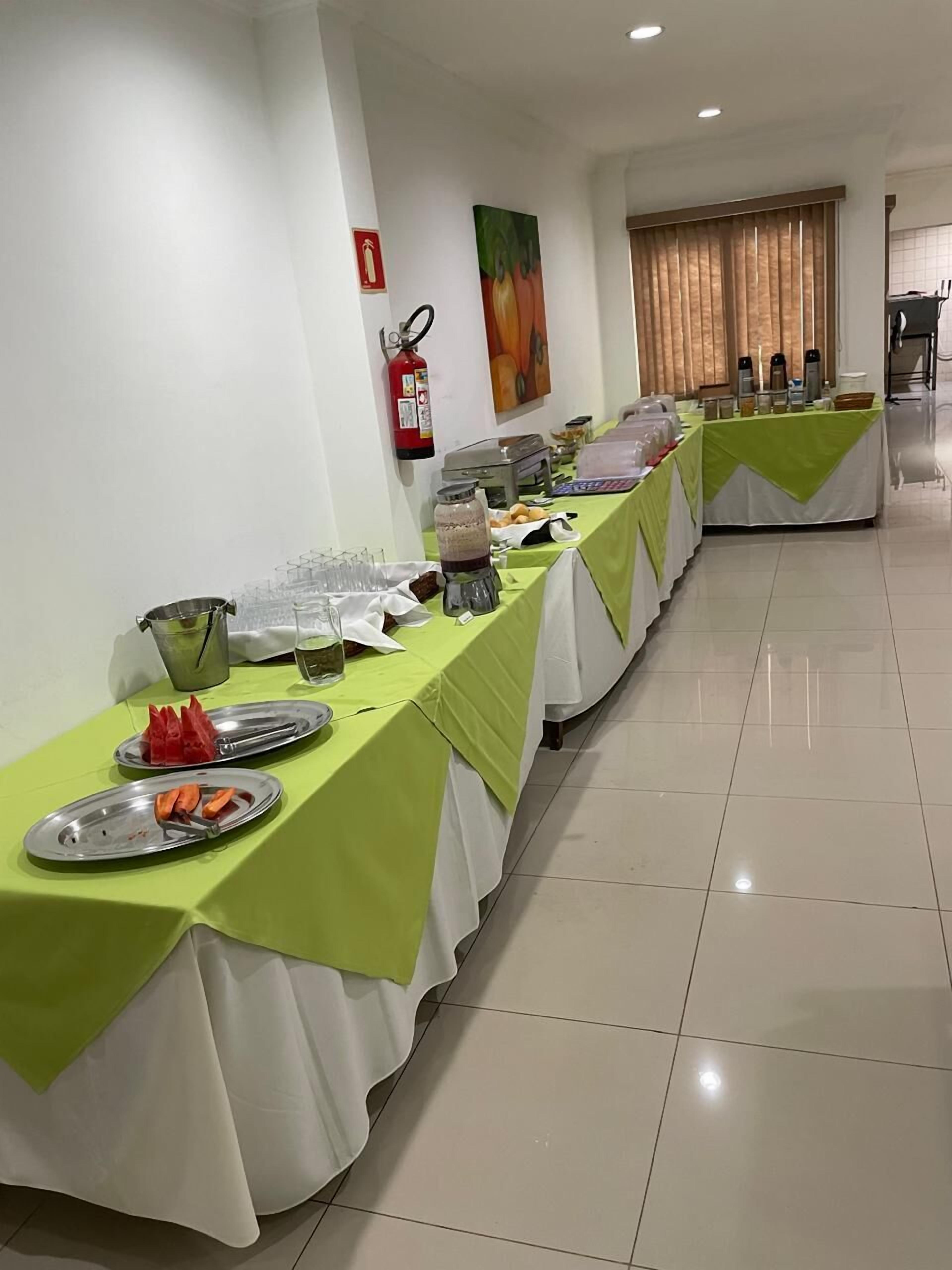 Uma longa mesa de buffet com toalhas de mesa verdes e brancas, diversos alimentos e um extintor de incêndio na parede.