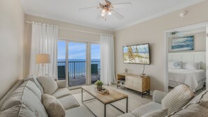 A spacious living room with a sofa, coffee table, and a ceiling fan. There is a balcony with a view of the ocean.