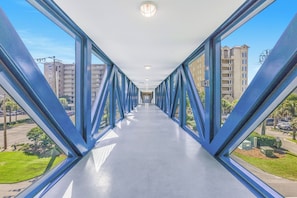 A corridor with glass walls and blue metal beams, leading to a building with multiple floors and balconies.