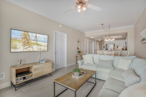 A modern living room with a sofa, coffee table, and TV. There is a dining area with a table and chairs in the background.
