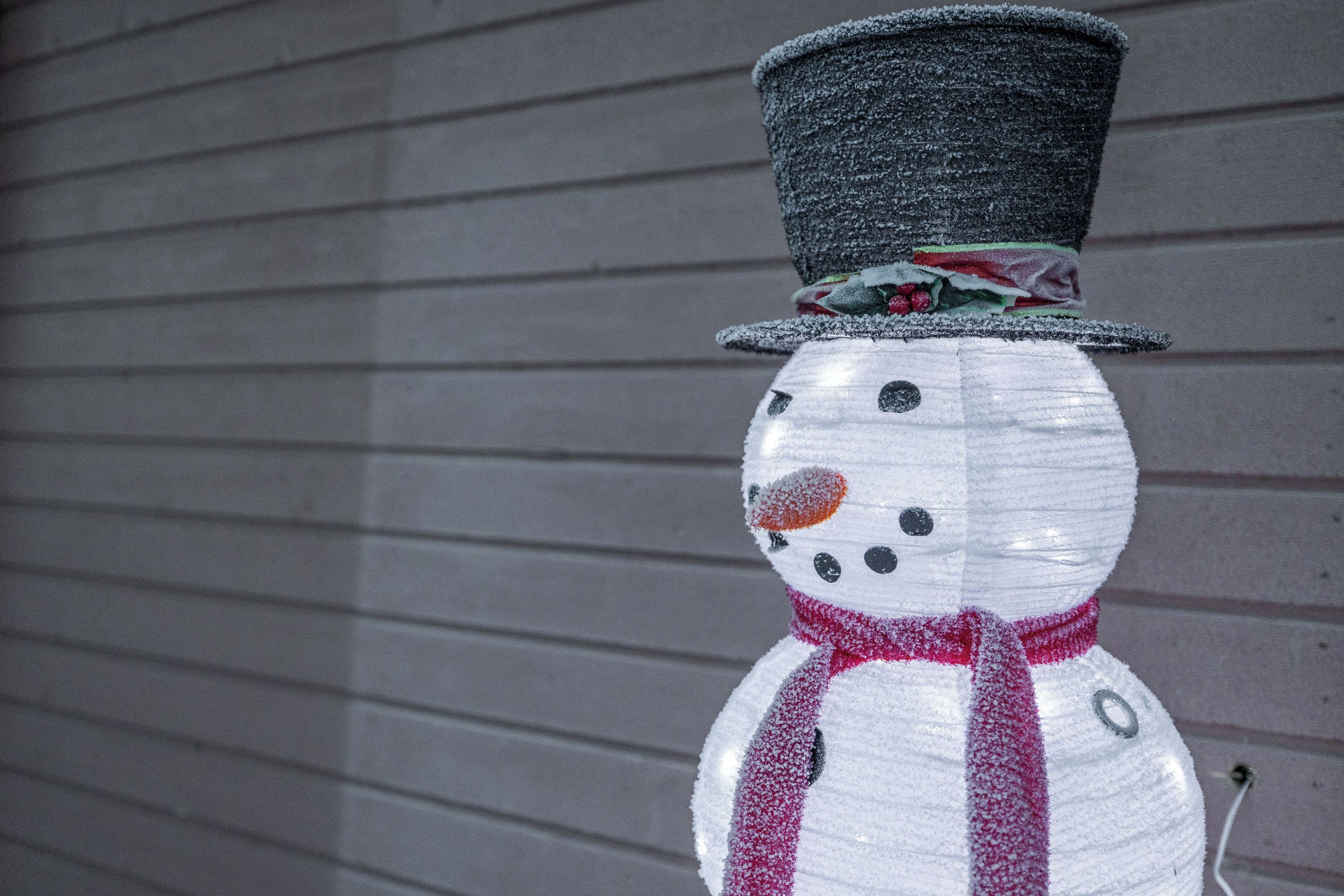 Un bonhomme de neige portant un haut-de-forme noir et une écharpe rouge, adossé à un mur gris.