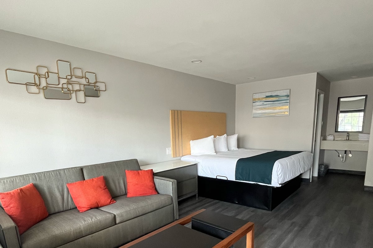 A modern hotel room with a grey sofa, a wooden coffee table, a bed with a green blanket, and a wall-mounted artwork.