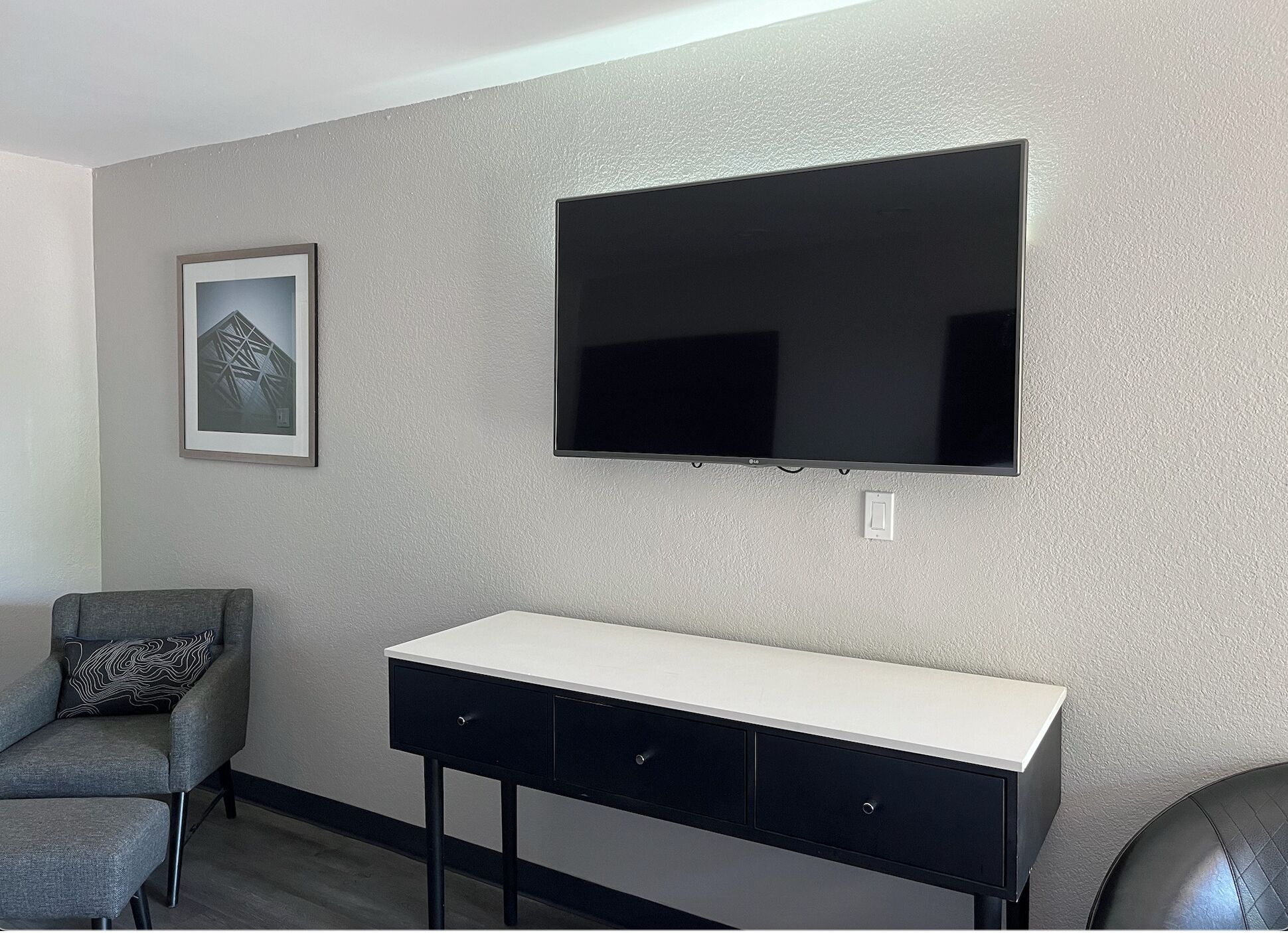 A modern room with a flat-screen TV mounted on the wall, a grey armchair with a patterned cushion, and a white desk with two drawers.