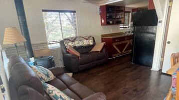A living room with a brown sofa, a patterned rug, and a kitchen area with wooden cabinets and a black refrigerator.