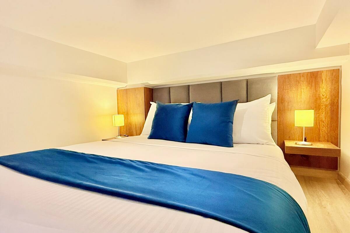 A hotel room with a large bed, two bedside tables with lamps, a wooden headboard, and a blue throw blanket.