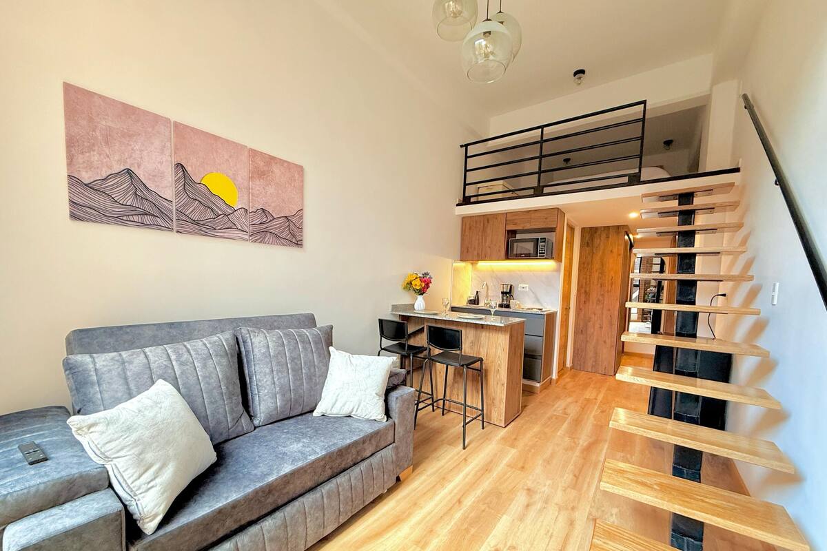 A modern living room with a grey sofa, wooden staircase, and a kitchen area with a bar.