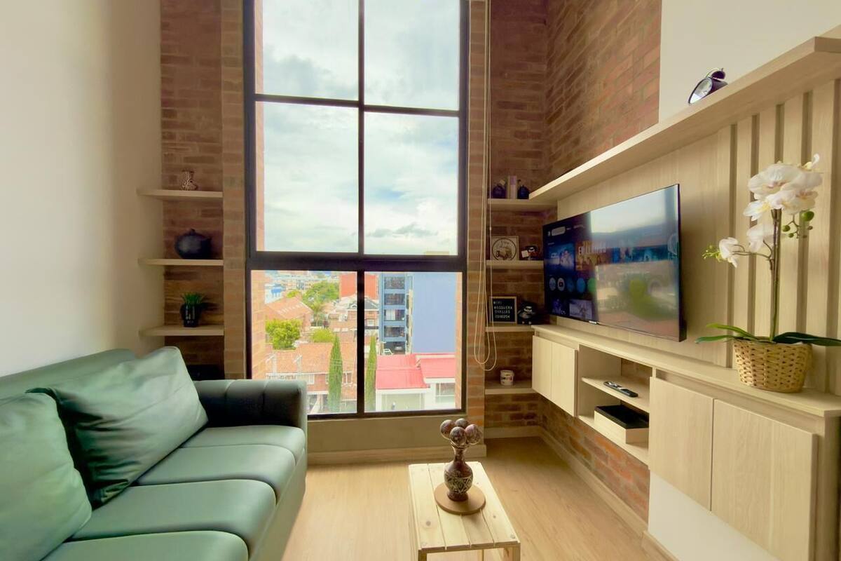 A modern living room with a green sofa, a TV on a wooden cabinet, and a large window with a view of buildings.