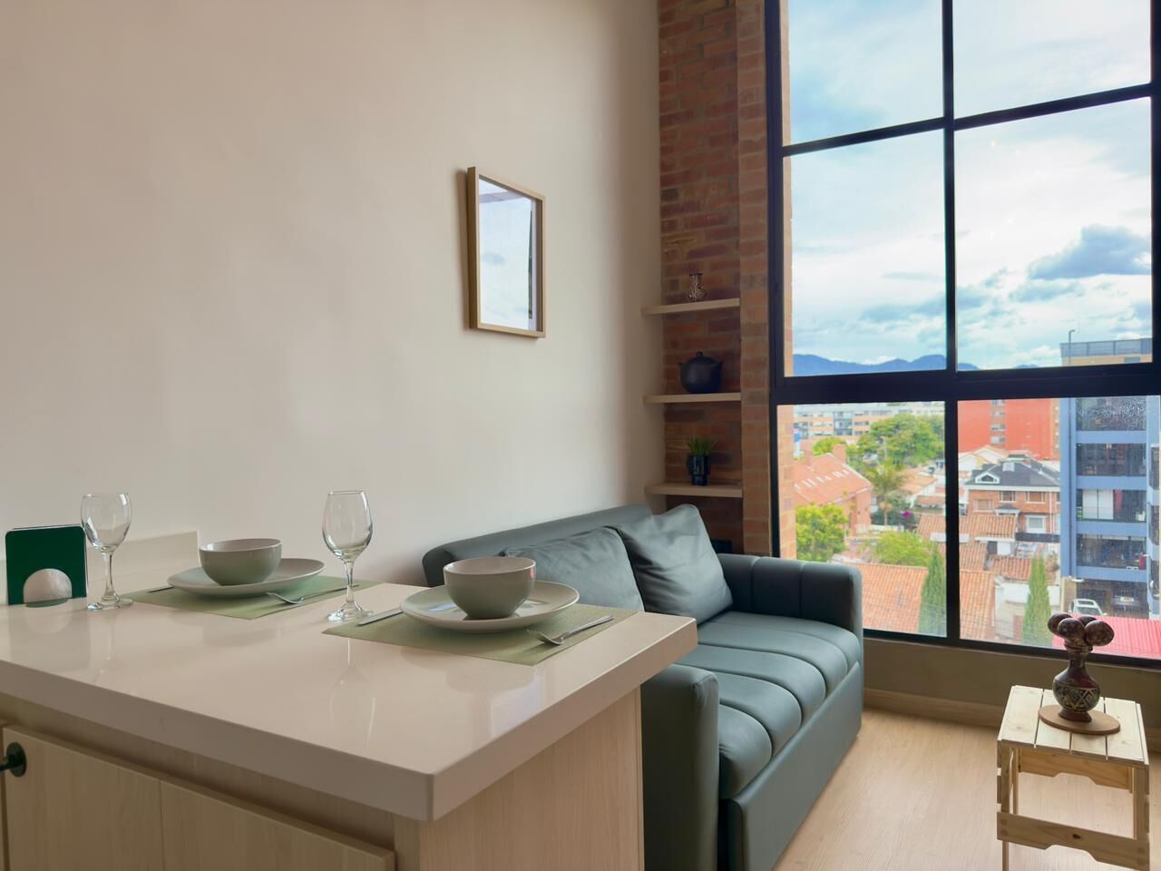 A modern living room with a dining area, a grey sofa, a large window with a city view, and a small table with a vase.