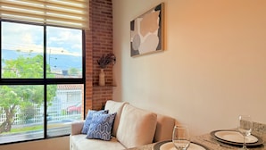 A modern living room with a large window, a sofa, a dining table set with plates and wine glasses, and a brick wall with a decorative vase. - Atelier Cedritos by Weeki. (Bogotá)