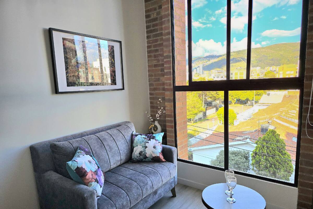 A room with a grey sofa, a framed picture on the wall, a vase with flowers, and a view of a landscape through a large window.