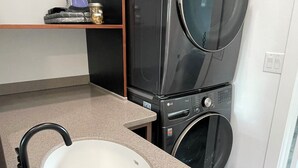 A compact laundry area with a washing machine and dryer stacked vertically, a sink with a faucet, and a shelf with folded towels. - Tiny Retreat at Stoney Creek  (Seward)