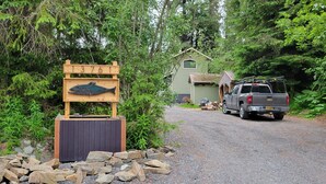 A wooden sign with the number 13761, a fish sculpture, and a small shed in a forested area with a gravel path and a truck parked nearby. - Tiny Retreat at Stoney Creek  (Seward)
