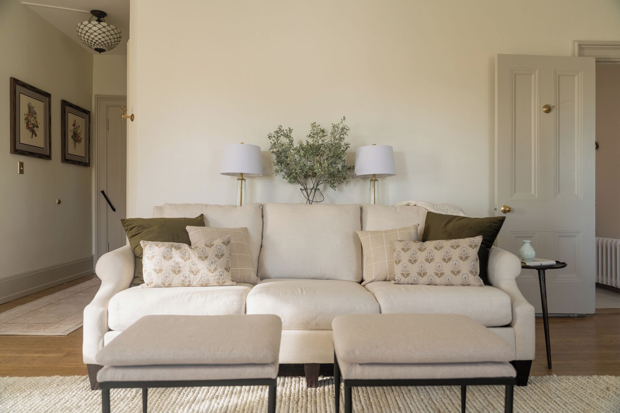 A living room with a beige sofa, two matching ottomans, a small side table, and two wall-mounted lamps.