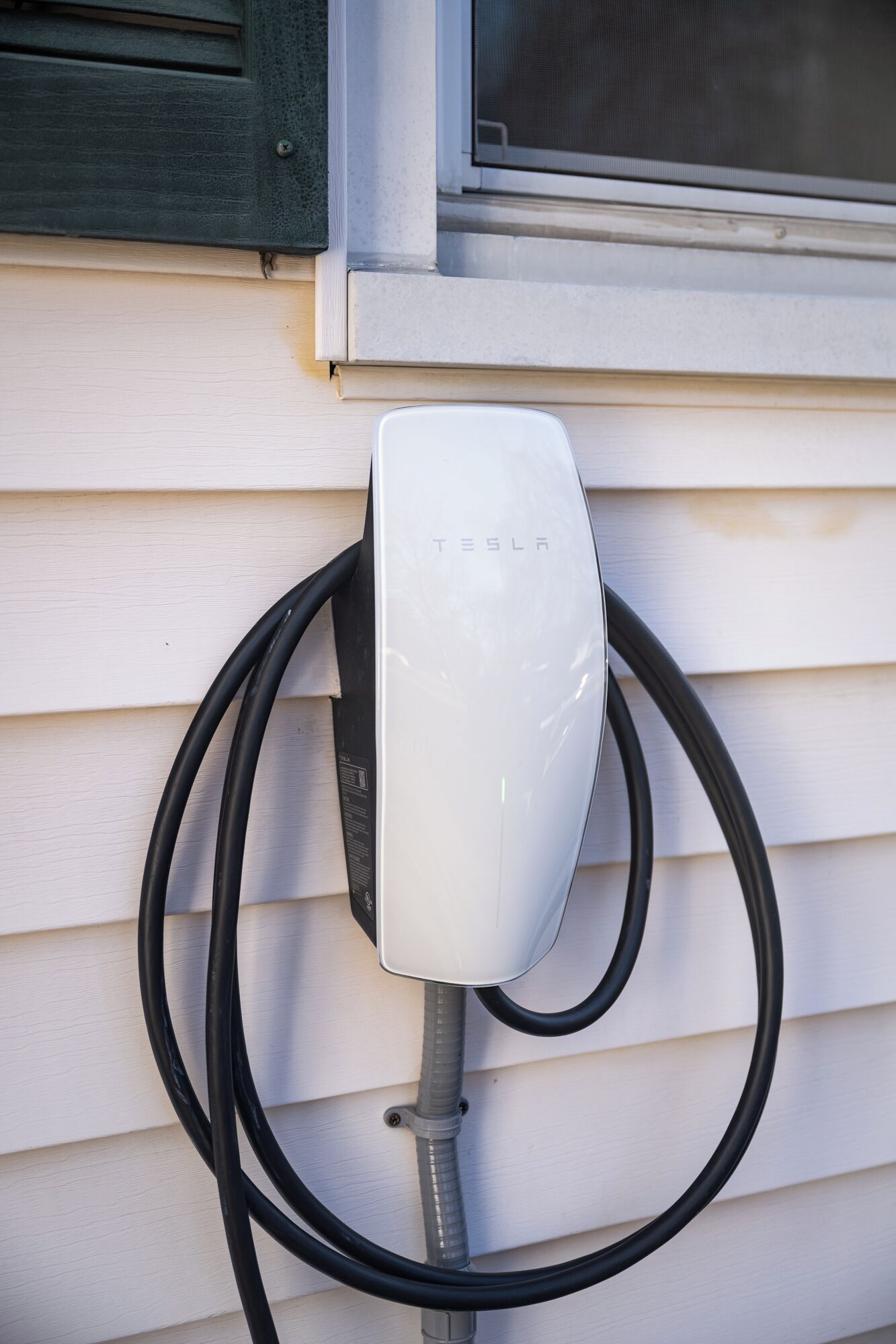 Een Tesla-laadstation gemonteerd aan een huis met beklede gevel en een raam erboven.