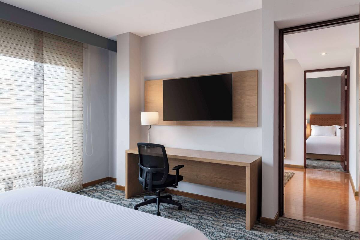 A hotel room with a bed, a desk with a chair, a television mounted on the wall, and a door leading to another room.