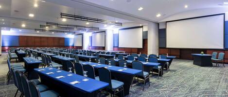 Un salón de conferencias con filas de mesas y sillas, una pantalla grande y un atril.