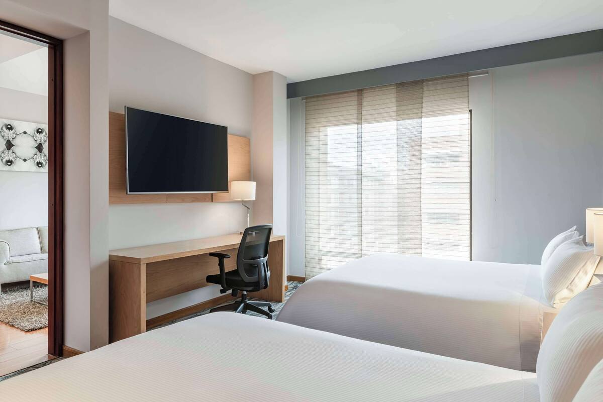 A hotel room with two beds, a desk, a chair, a TV, and a window with blinds.