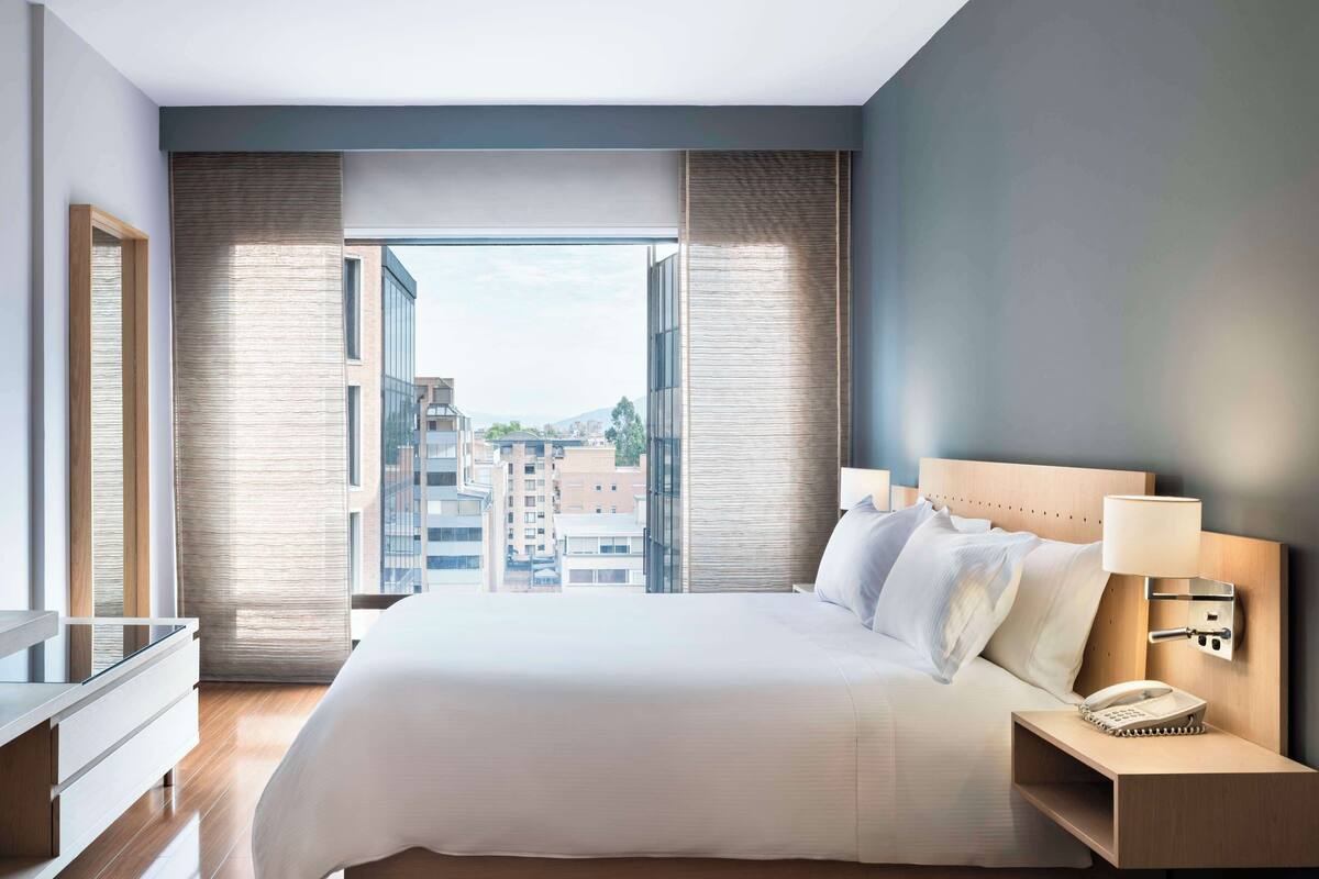 A modern hotel room with a large bed, bedside table, and a view of the cityscape through floor-to-ceiling windows.