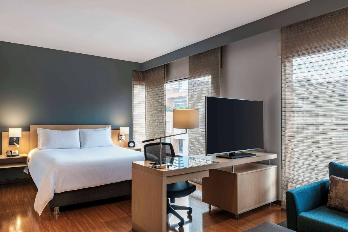 A hotel room with a bed, desk, chair, TV, lamp, and window with blinds.