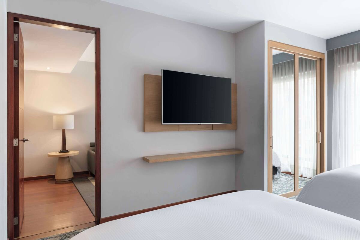 A hotel room with a bed, a mounted TV, a small table with a lamp, and a sliding glass door leading to a balcony.