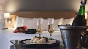 Un lit avec du linge de lit blanc, une table de chevet avec une lampe, une bouteille de champagne, deux coupes de champagne et une assiette avec des aliments.