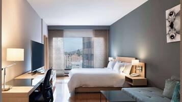 A modern hotel room with a large bed, a desk with a computer, a sofa, and a view of the city through large windows.