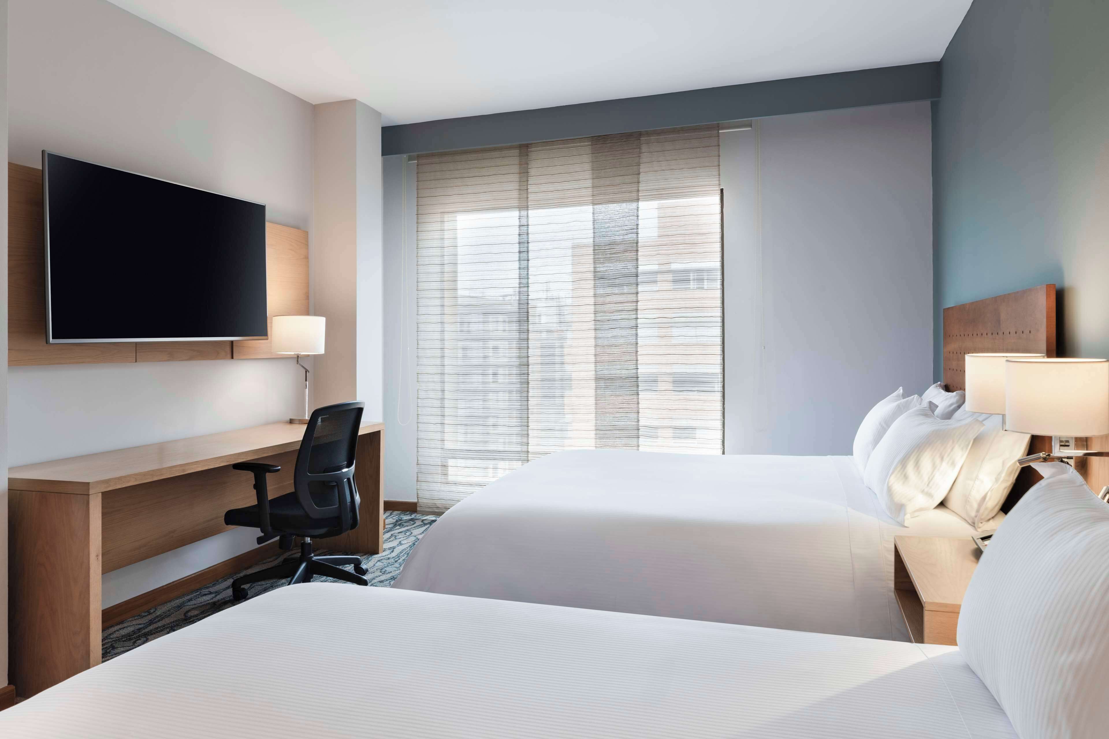 Habitación de hotel con dos camas, un escritorio con silla, un televisor de pantalla plana y una ventana con persianas.